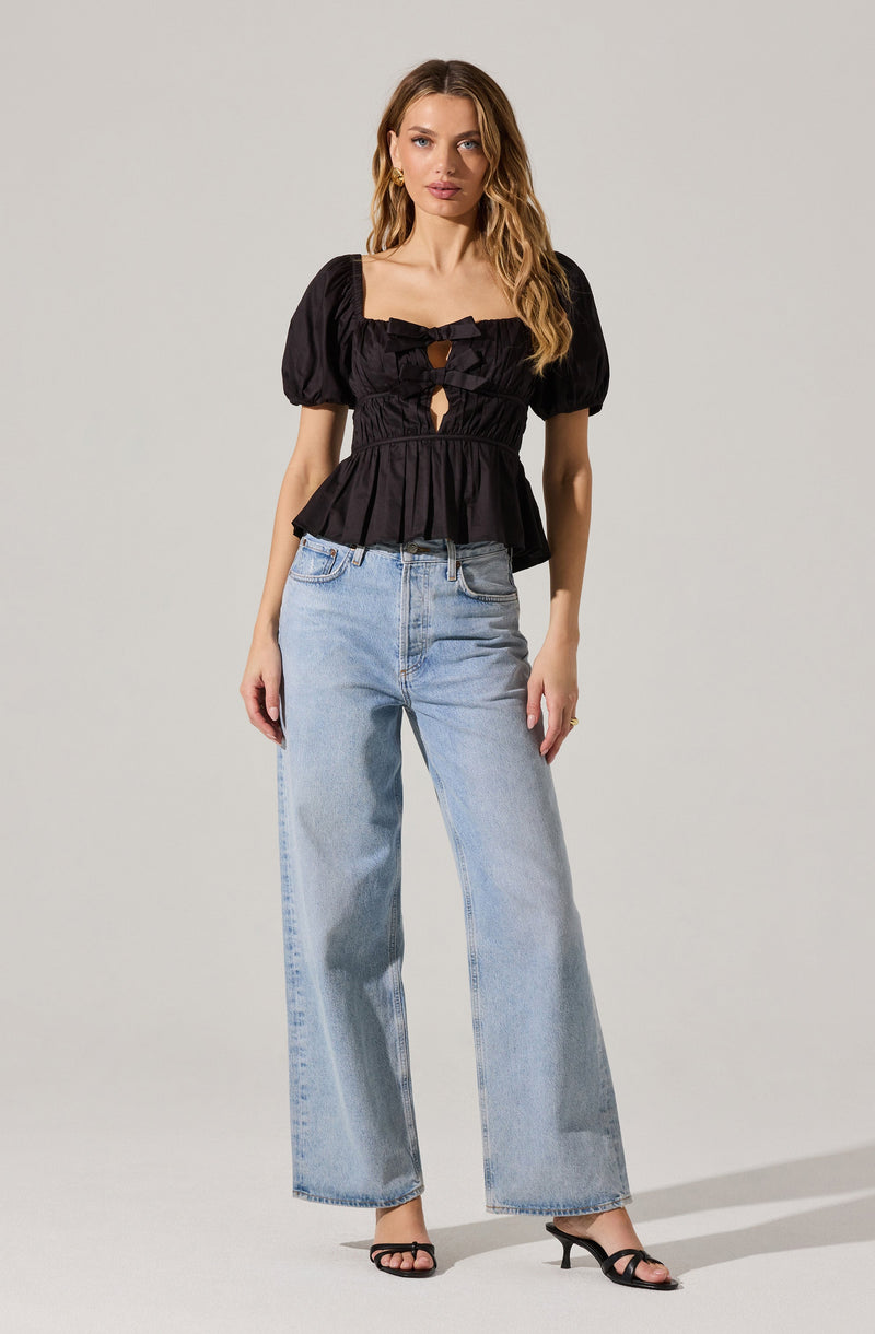 A woman is wearing a black, short-sleeved top with puffed sleeves and a square neckline. The top features a decorative bow at the center of the chest and cut-out details along the front. She is also wearing light-wash, high-waisted wide-leg jeans and black open-toe high-heeled sandals.