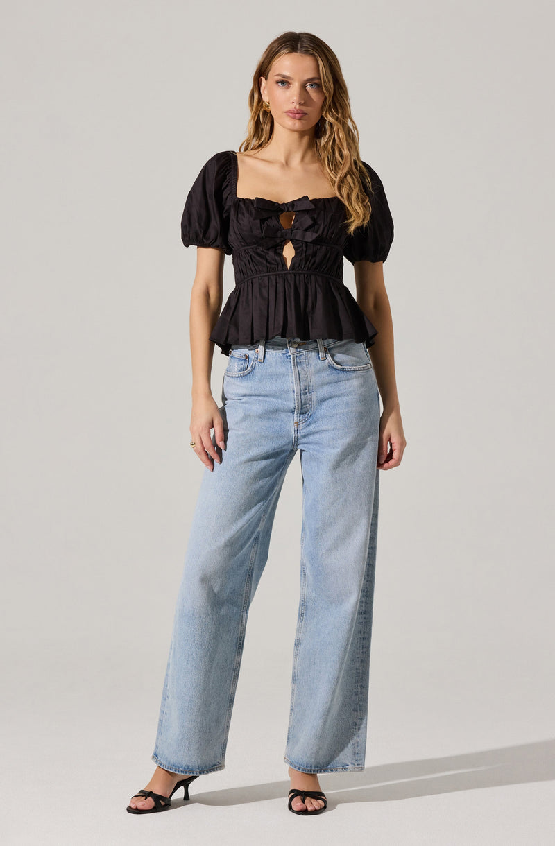A woman is wearing a black, short-sleeved top with puffed sleeves and a square neckline, featuring a keyhole cutout at the bust with a bow tie detail. She pairs it with high-waisted, light-wash wide-leg jeans and black high-heeled sandals.