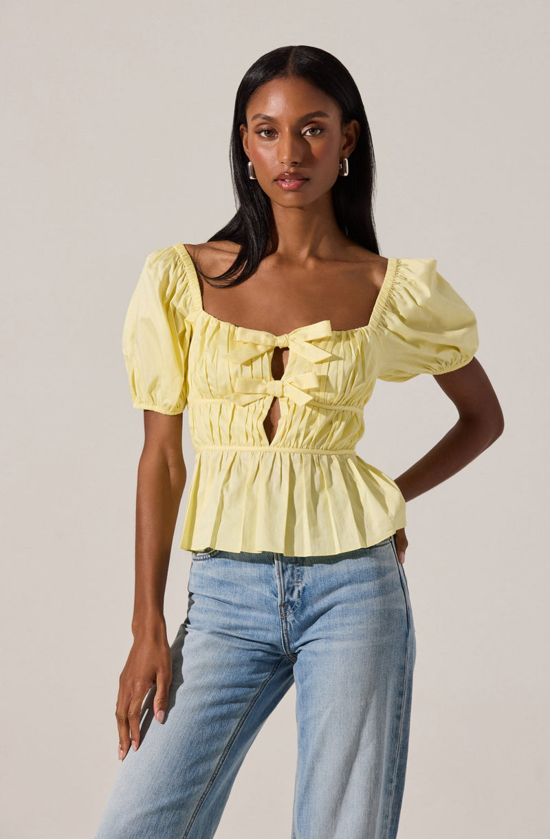 A woman is wearing a yellow top with puffed short sleeves, a square neckline, and a front tie detail with cut-out openings. The top has a gathered, ruffled hem that falls at the waist. She is also wearing light blue high-waisted jeans.