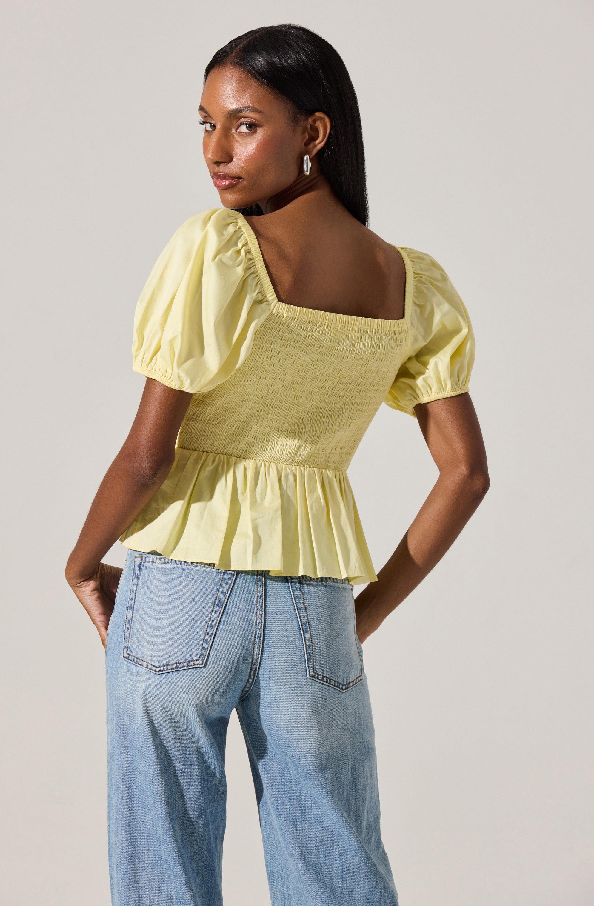 A woman is wearing a yellow top with puffed short sleeves, a square neckline, smocking at the bodice, and a ruffled peplum hem. The top features gathered fabric at the shoulders and a fitted, textured waistline. She is also wearing light-wash jeans with a relaxed fit, featuring visible back pockets and stitched detailing. The background is plain and light-colored.