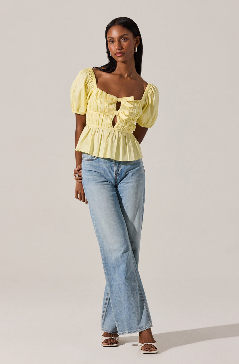 A woman is wearing a light yellow, short-sleeved blouse with puffed sleeves and a square neckline. The blouse features gathered fabric with two front keyhole cutouts, tied with bows, and has a ruffled hem. She pairs it with light-wash, high-waisted, wide-leg jeans that reach the floor. She is also wearing open-toe heels.