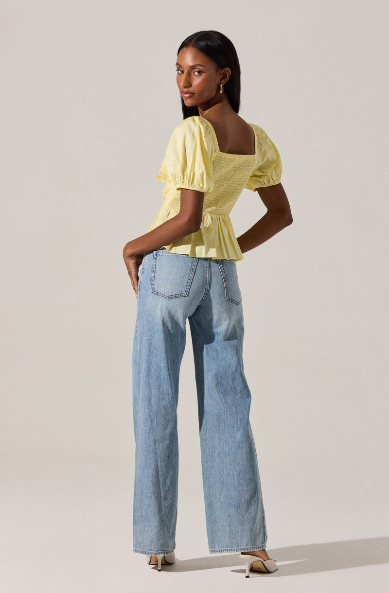 A woman is wearing a yellow top with short, puffed sleeves that have gathered cuffs. The top has a square neckline, a fitted bodice with smocking detail, and a ruffled hem that flares out slightly. She is also wearing light blue, high-waisted, wide-leg jeans with back pockets, paired with beige pointed-toe heels.