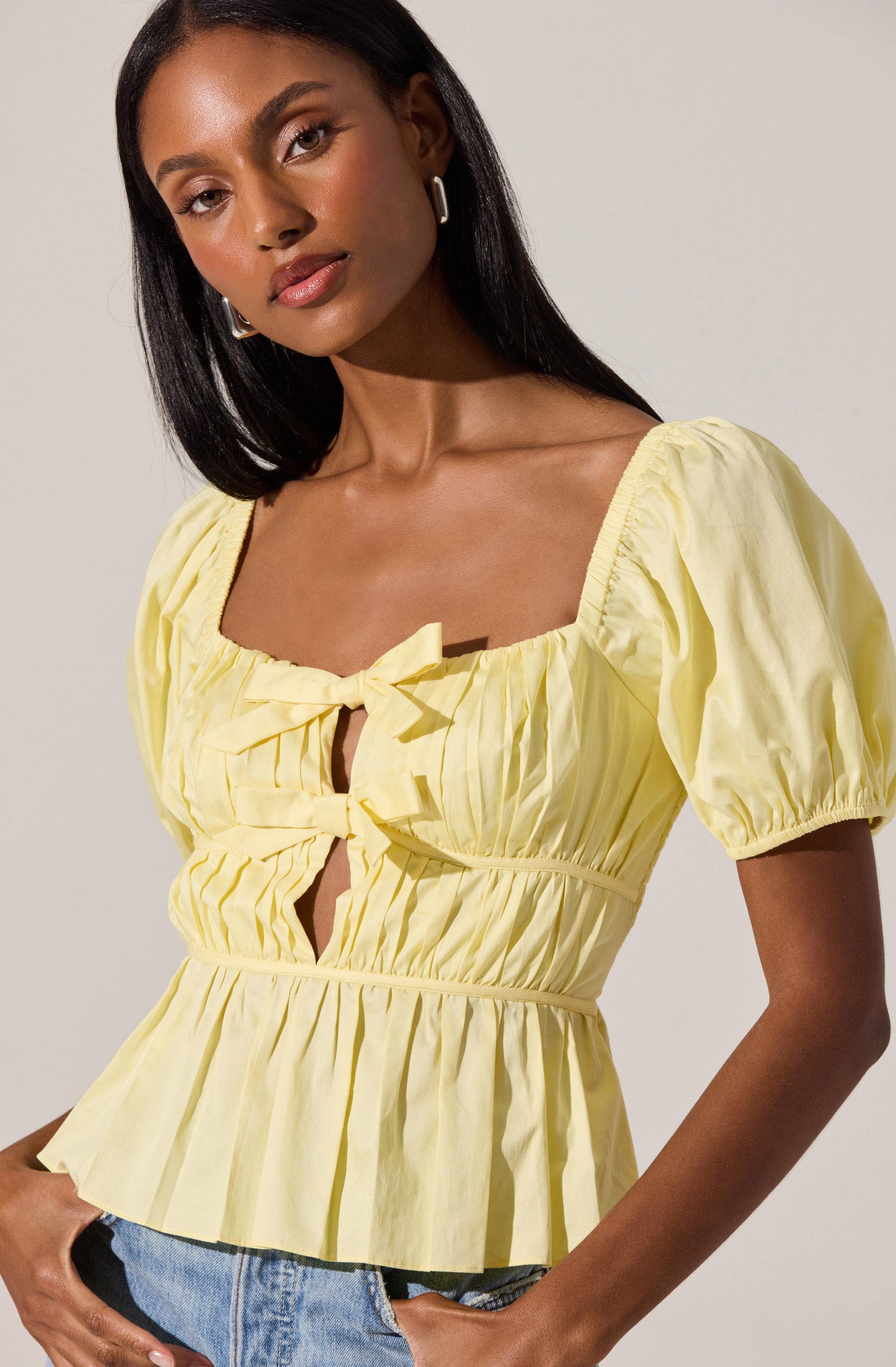 A woman is wearing a light yellow, short-sleeved blouse with a square neckline and puffed sleeves. The blouse features a front tie detail at the neckline, pleats along the bodice, and a gathered waist that creates a peplum effect. She is also wearing light blue denim jeans.