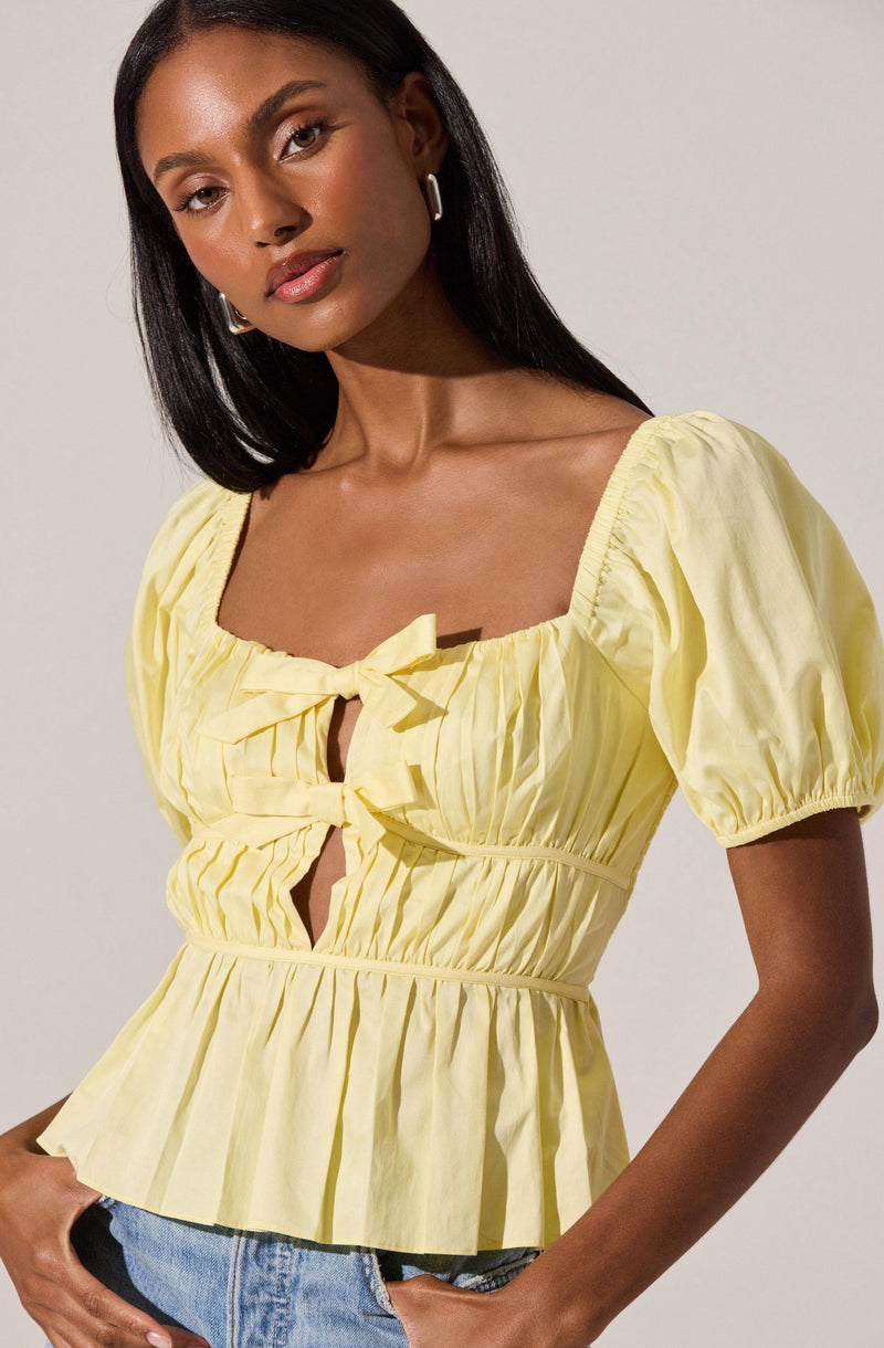 A woman is wearing a light yellow, short-sleeved blouse with a square neckline and puffed sleeves. The blouse features a front tie detail at the neckline, pleats along the bodice, and a gathered waist that creates a peplum effect. She is also wearing light blue denim jeans.