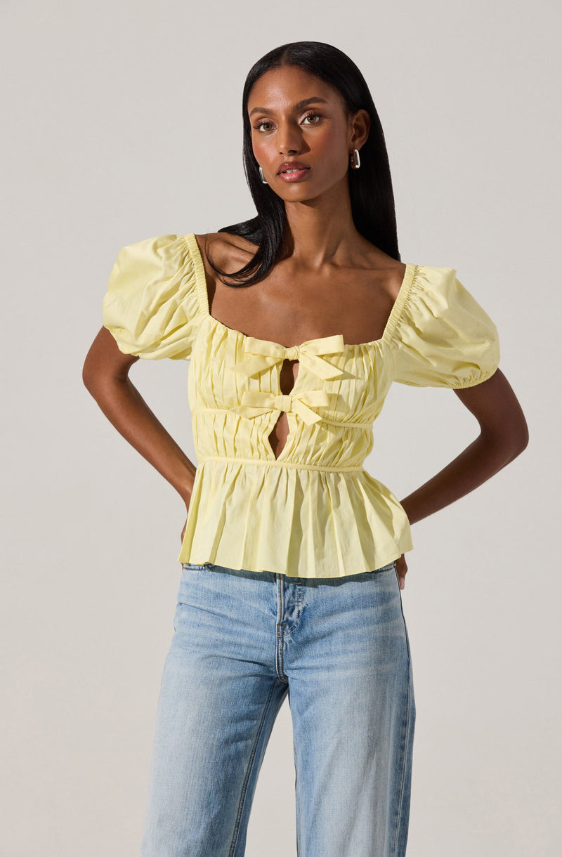 The woman is wearing a light yellow blouse with puffed short sleeves, a square neckline, and decorative bows with cut-out details on the front. She is also wearing light-wash blue jeans.