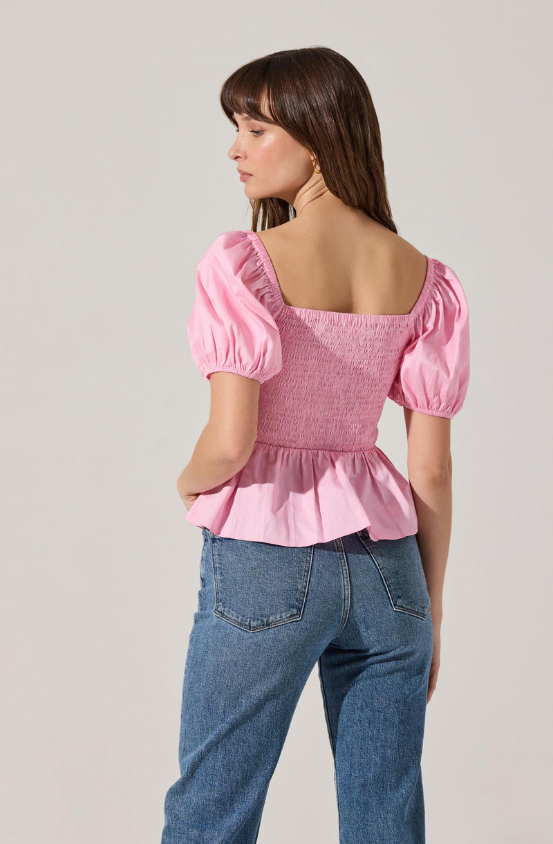 A woman is wearing a pink puff-sleeve top with a square neckline and a gathered, smocked bodice. The top has a loose, ruffled hem that extends below the waist. She pairs it with high-waisted blue jeans.