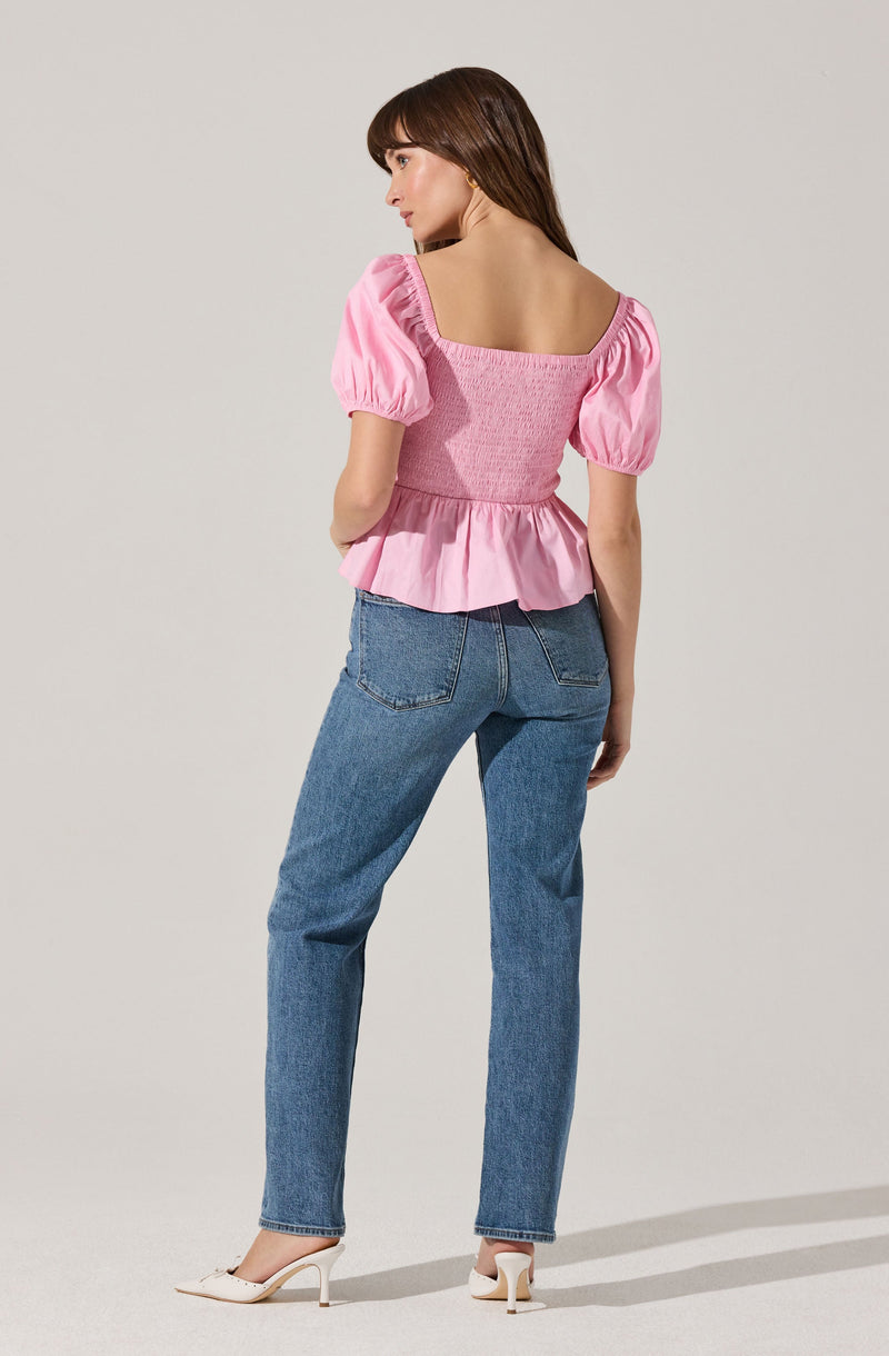 A woman is wearing a pink top with puffed short sleeves, a square neckline, and a ruffled hem that flares out at the waist. She pairs it with high-waisted blue jeans and white high-heeled shoes.