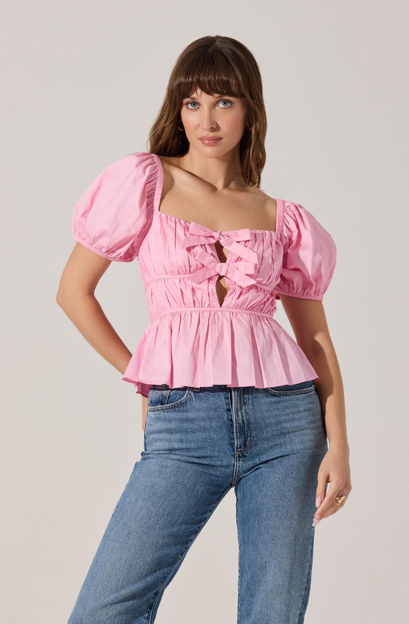 A woman is wearing a pink blouse with puffed short sleeves, a square neckline, and a ruched, gathered waist. The blouse features a decorative bow and cut-out detail at the front. She is paired with light blue high-waisted jeans.