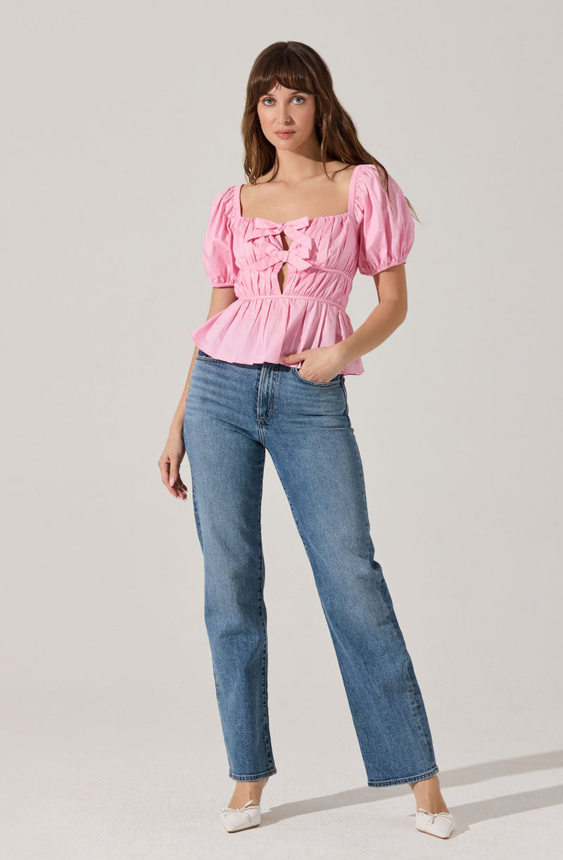 A woman is wearing a light pink, short-sleeved top with puffed sleeves and a square neckline. The top features shirred fabric and decorative bow details at the front, with a peplum-style ruffled hem. She is also wearing high-waisted, straight-leg blue jeans and white shoes.