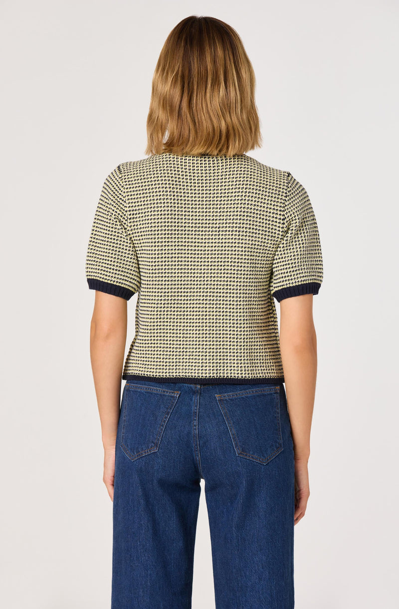 A woman is wearing a short-sleeved, textured beige and black patterned top with dark contrasting trim on the sleeves and hem, paired with high-waisted blue jeans. The back view shows the fit and design of the clothing.