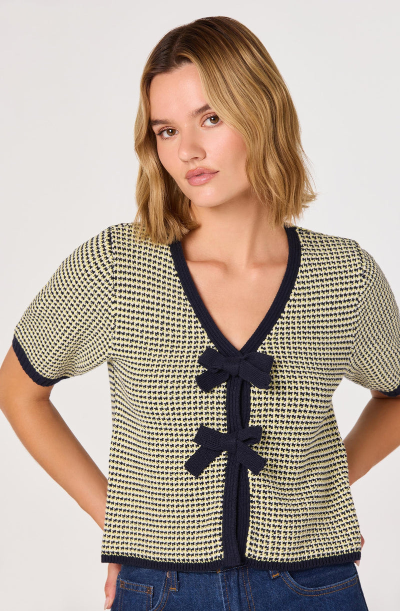 A woman is wearing a short-sleeved, V-neck sweater with a small, repeating geometric pattern in yellow and navy. The sweater features navy trim around the collar, sleeve cuffs, and down the front, accented with three matching navy bows along the front.