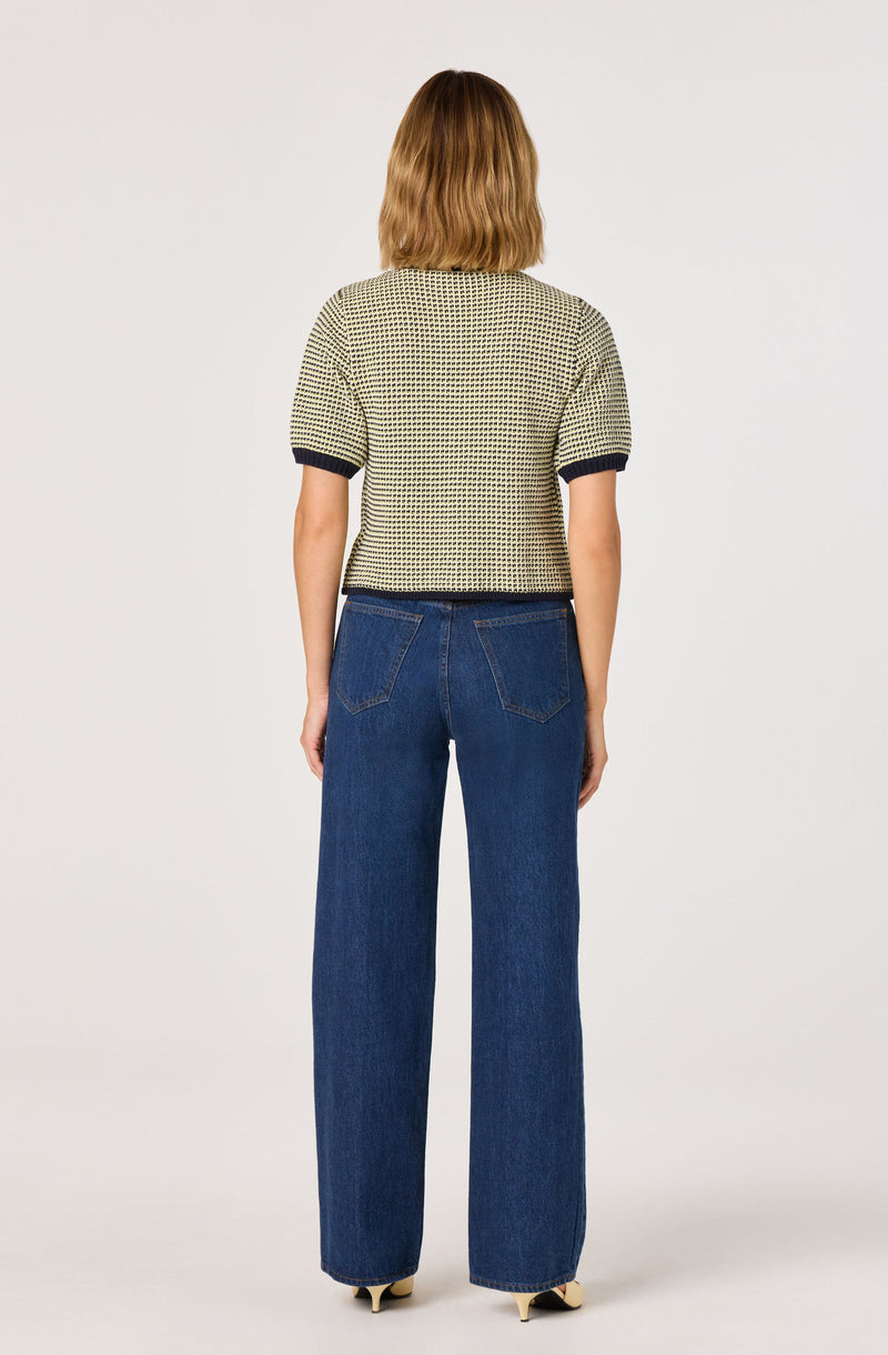 A person is wearing a short-sleeved, knitted sweater with a small checkered pattern in neutral tones and dark trim at the sleeves. They are also wearing high-waisted, wide-leg blue jeans. The outfit is casual, with the sweater and jeans paired together.