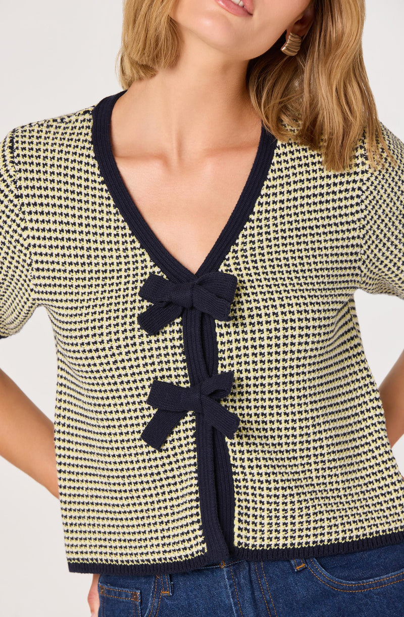 A woman is wearing a short-sleeved, knitted cardigan with a yellow and black houndstooth pattern. The cardigan has a navy blue trim around the neckline and sleeve edges, and features two navy blue bows attached to the front opening.