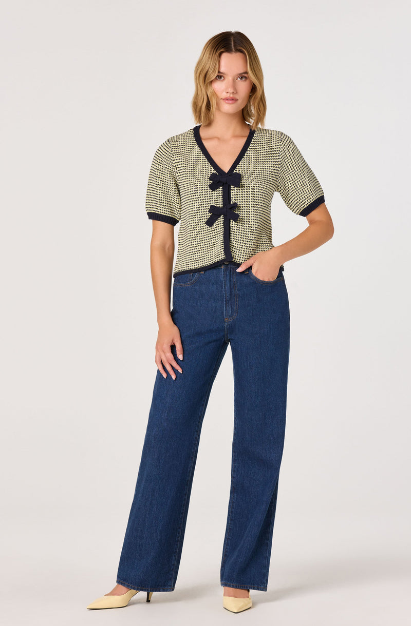 A woman is wearing a short-sleeved, V-neck cardigan with a small checkered pattern and navy trim along the neckline, sleeve edges, and front opening. The cardigan has two navy bows on the front. She pairs it with high-waisted, wide-leg blue jeans and beige pointed-toe heels.