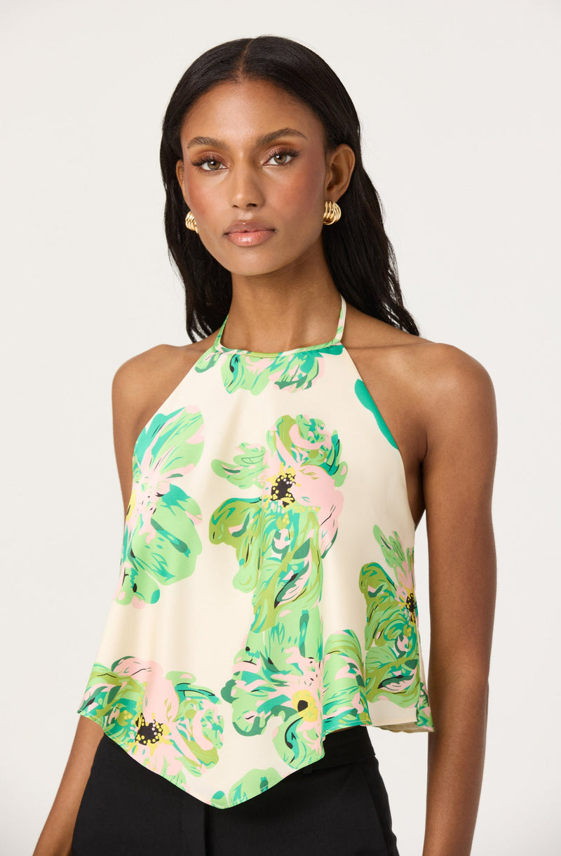 A woman is wearing a sleeveless halter top with a loose, flowy fit. The top features a high neckline with thin straps and is decorated with a colorful floral pattern in shades of green, pink, and yellow on a light cream background.