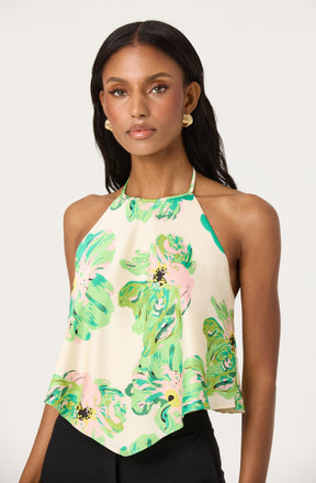 A woman is wearing a sleeveless halter top with a loose, flowy fit. The top features a high neckline with thin straps and is decorated with a colorful floral pattern in shades of green, pink, and yellow on a light cream background.