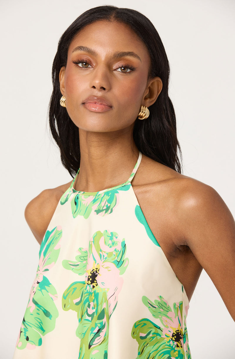 A woman is wearing a sleeveless, halter-neck dress with a floral pattern in shades of green, pink, and black on a light-colored background.