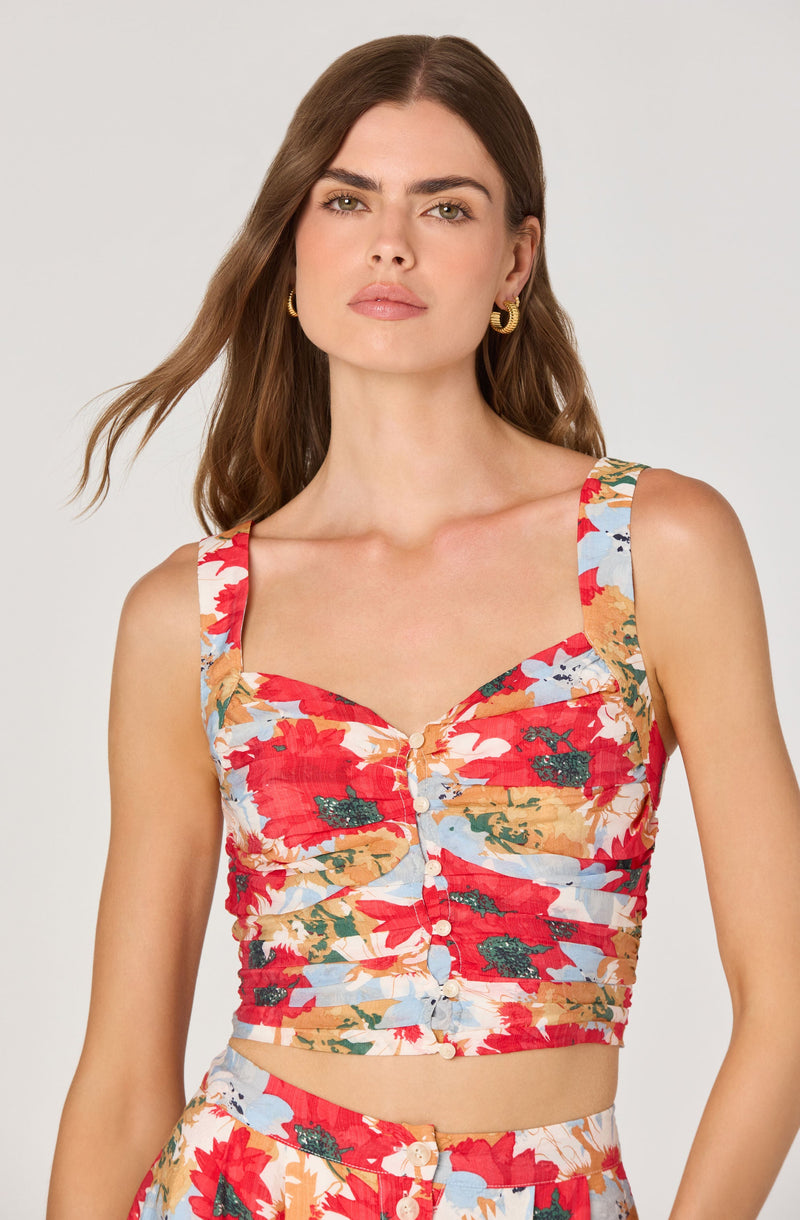 A woman is wearing a sleeveless, fitted crop top with wide straps. The top features a colorful floral pattern with large red flowers, green leaves, and light blue accents. The top has a button-down front with small white buttons running down the center.