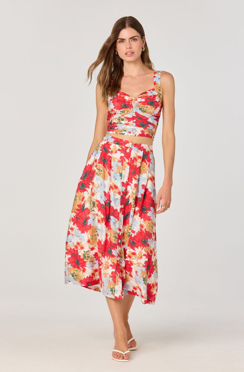 A woman is wearing a two-piece floral outfit consisting of a fitted crop top with wide shoulder straps and a high-waisted, flowy midi skirt. The fabric features a vibrant, multicolored floral print with red, yellow, white, and green flowers. She is also wearing white open-toe heels and small hoop earrings.
