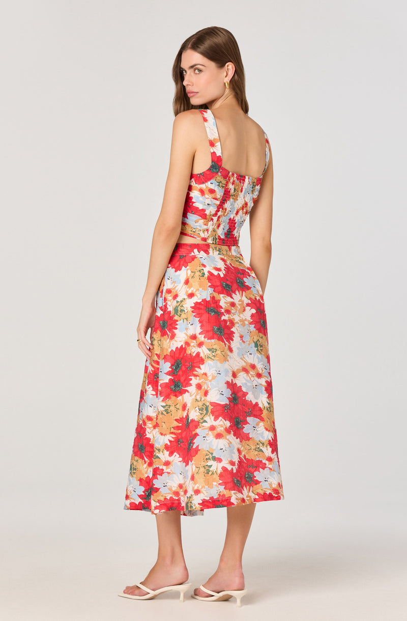 A woman is wearing a sleeveless, fitted crop top and a matching high-waisted, ankle-length skirt. Both pieces feature a vibrant, multicolored floral pattern with shades of red, pink, orange, yellow, and light blue. She is also wearing light-colored, open-toe high-heeled sandals.