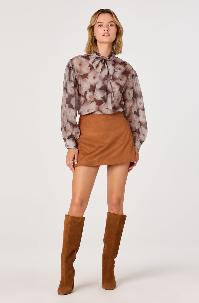 A woman is wearing a semi-sheer blouse with a floral print and a large bow at the collar, along with a high-waisted, tan suede mini skirt. She completes her outfit with knee-high tan suede boots.
