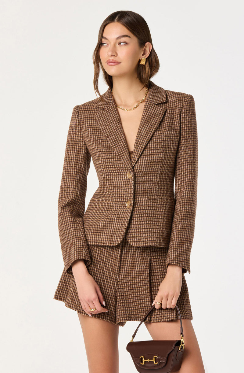 A woman is wearing a tailored, fitted blazer and a matching pleated mini skirt, both made from a brown and black houndstooth patterned fabric. The blazer has a notched lapel collar, long sleeves, and two buttons at the front. She is accessorized with gold hoop earrings, a gold chain necklace, and rings on her fingers. She is holding a small brown handbag with a gold buckle and a short strap.