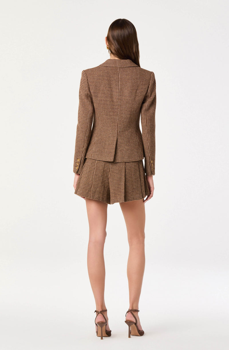 A woman is wearing a tailored, checked blazer in shades of brown and beige, paired with matching high-waisted shorts featuring pleats. She is also wearing tan high-heeled sandals with ankle straps.