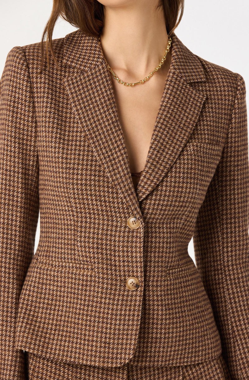 A woman is wearing a fitted brown houndstooth blazer with a notched lapel collar and two large buttons. The blazer has structured shoulders and a tailored silhouette. She is also wearing a gold chain necklace.