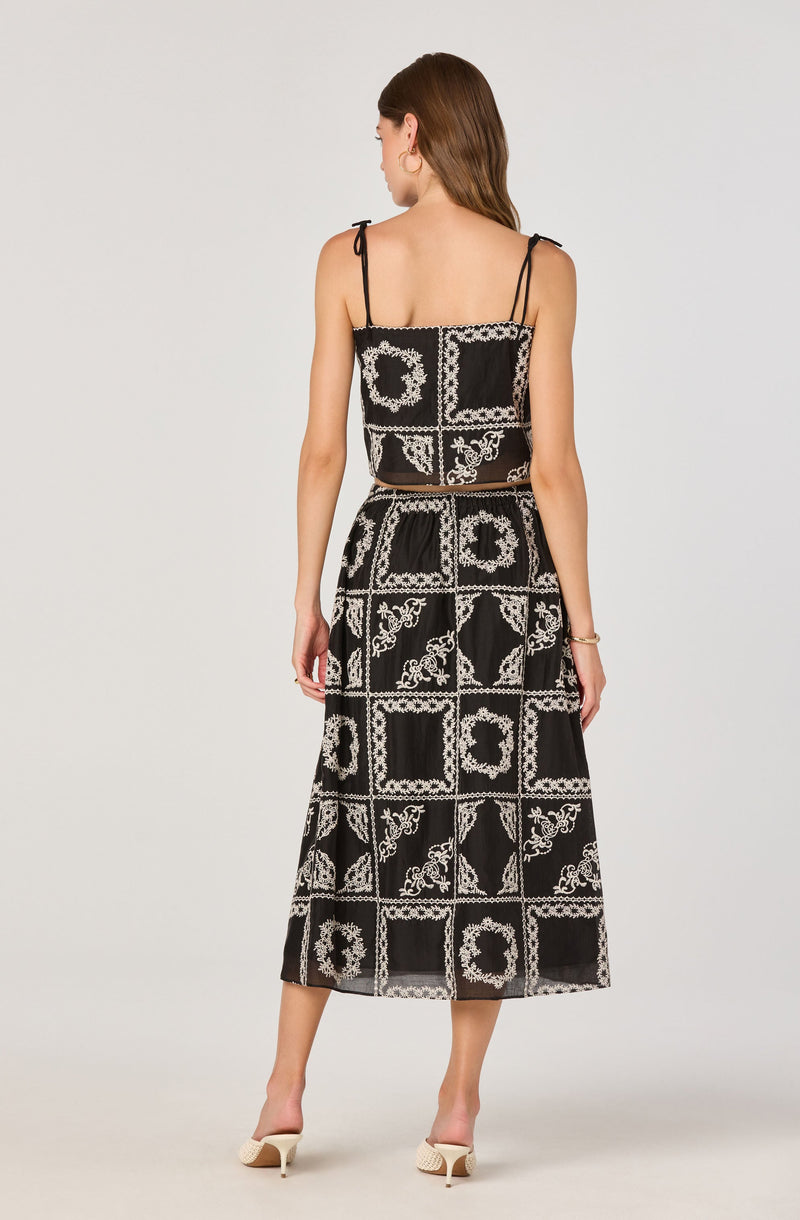 A woman is wearing a black sleeveless dress with thin shoulder straps. The dress features a pattern of white ornate floral and geometric designs arranged in square patches throughout the fabric. The top part is a slightly cropped tank style, and the skirt is long, reaching just above her ankles. She is also wearing light-colored high-heeled shoes.
