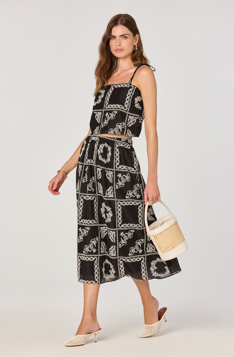 A woman is wearing a black two-piece outfit consisting of a spaghetti strap crop top and a matching midi skirt. The fabric features a white, intricate, bandana-inspired pattern arranged in a grid formation. She accessorizes with gold hoop earrings and a gold bracelet, and carries a white woven bucket bag. She is wearing open-toe, beige knit high-heeled mules.