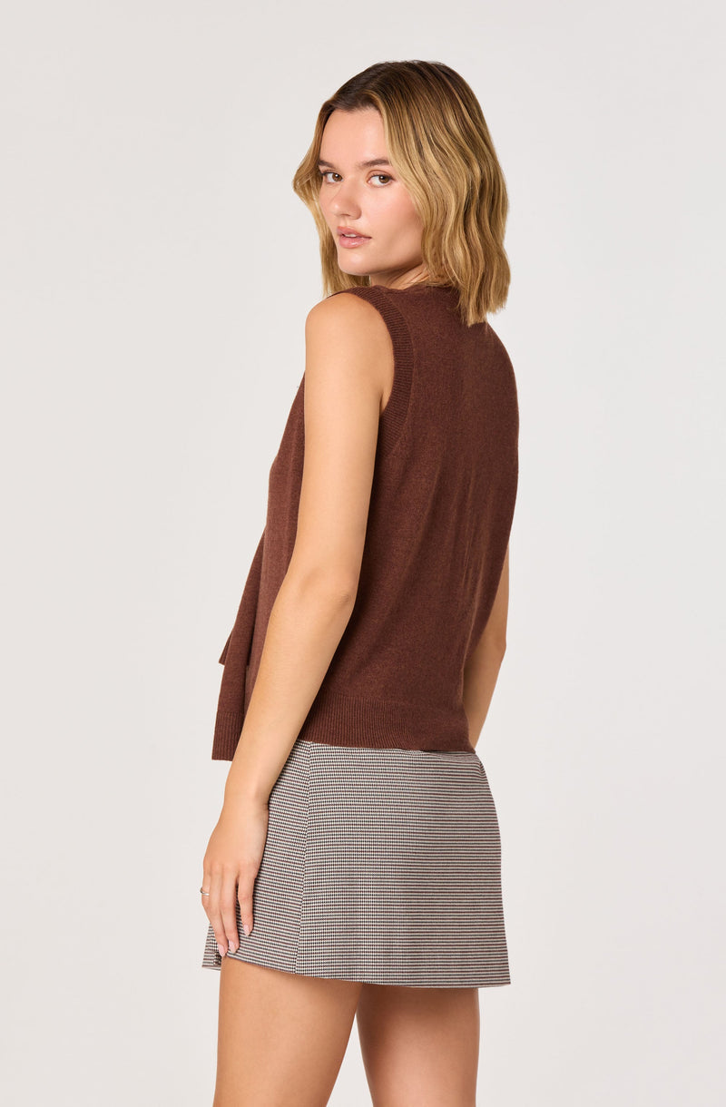 A young woman with shoulder-length wavy blonde hair is wearing a sleeveless, high-neck brown vest and a short, fitted skirt with a small checkered pattern in neutral tones.