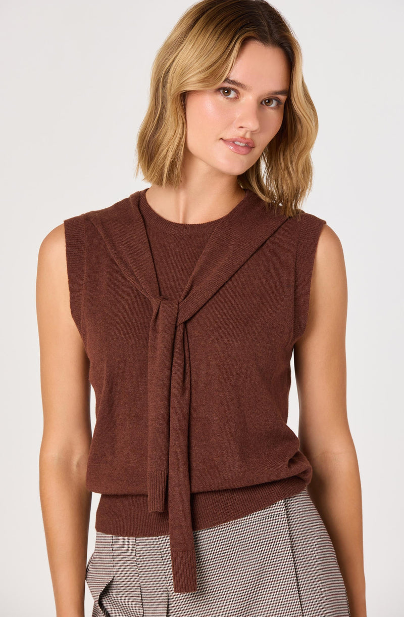 The woman is wearing a sleeveless, dark brown knitted top with a matching knitted tie at the neck. She pairs it with high-waisted, light-colored checkered trousers.