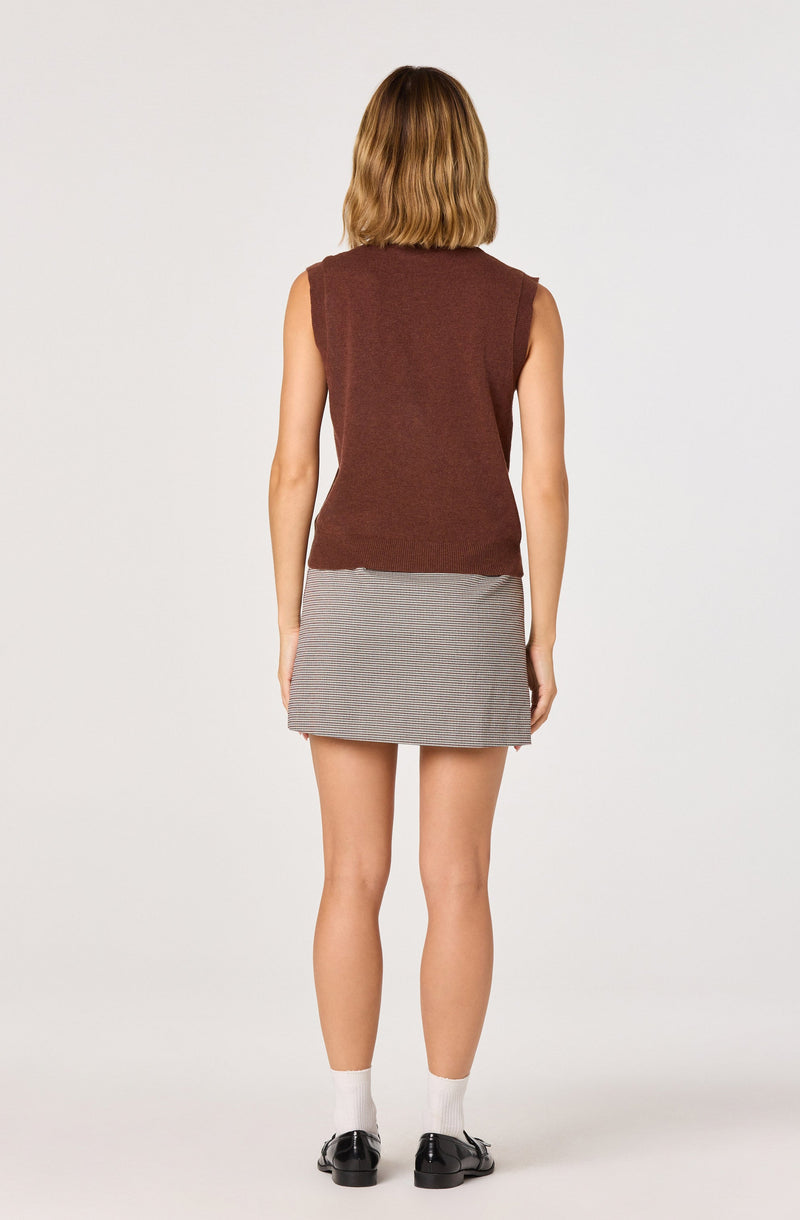 A woman is wearing a sleeveless, brown knit vest paired with a short, gray and white checkered skirt. She has on white socks and black shoes. Her hair is shoulder-length and light brown.