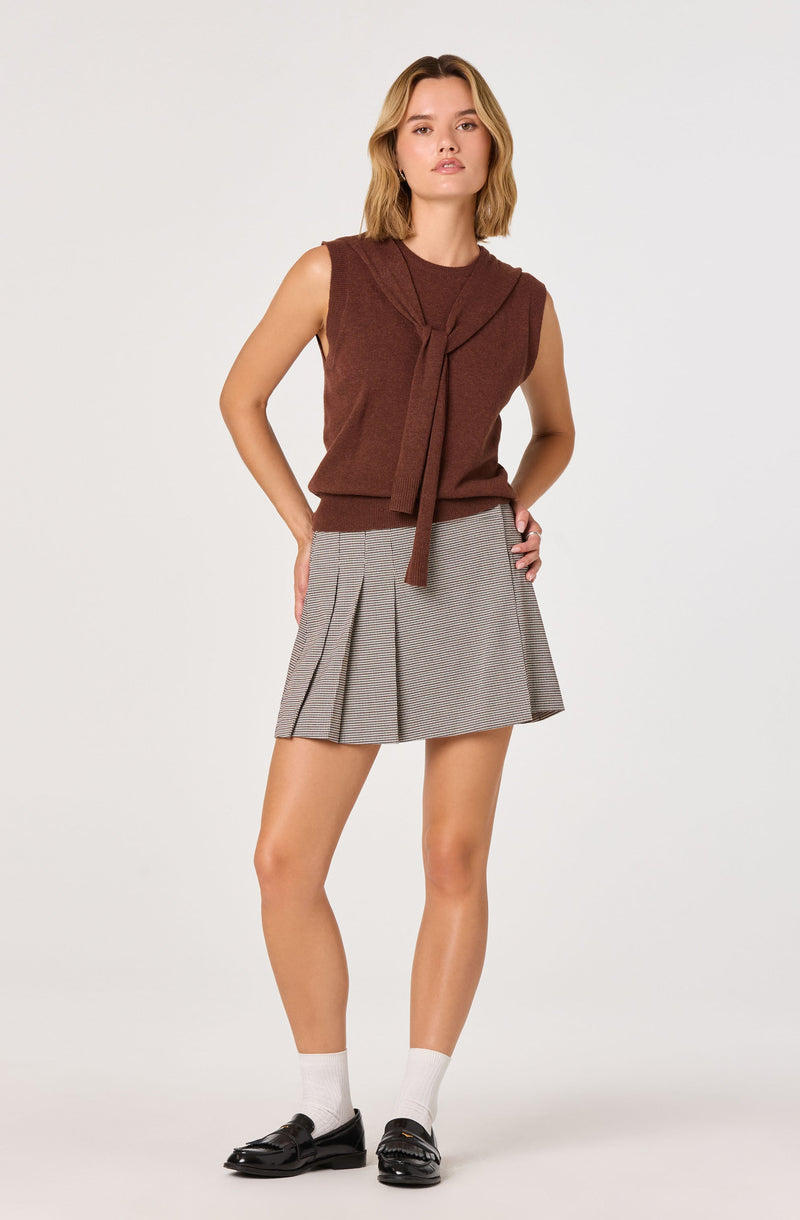 A young woman is wearing a sleeveless, brown knit top with a tied scarf detail at the neckline, paired with a high-waisted, pleated, gray plaid skirt. She has on white socks and black loafers.