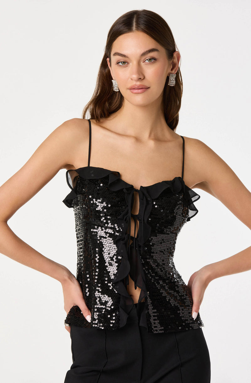 A woman is wearing a sleeveless black top with thin spaghetti straps. The top is decorated with shiny black and silver sequins and features ruffled trim along the neckline and down the front, with a ribbon tie detail at the center. She is also wearing large, square, silver earrings.