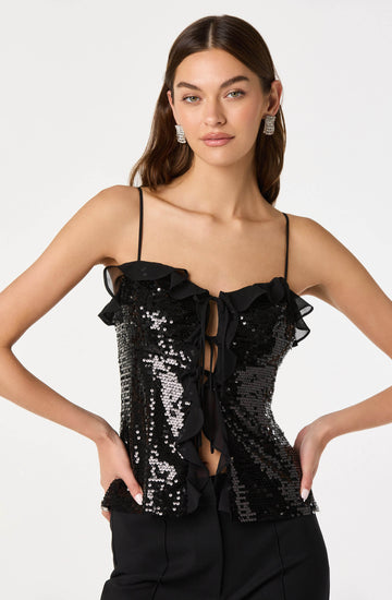 A woman is wearing a sleeveless black top with thin spaghetti straps. The top is decorated with shiny black and silver sequins and features ruffled trim along the neckline and down the front, with a ribbon tie detail at the center. She is also wearing large, square, silver earrings.
