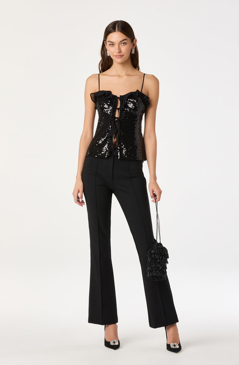A woman is wearing a black sequined camisole top with thin spaghetti straps, featuring ruffle details at the neckline and a front lace-up detail. She pairs it with high-waisted black tailored pants that have a slight flare at the bottom. She is holding a small black handbag with fringe details and is wearing black and white pointed-toe heels with embellishments.