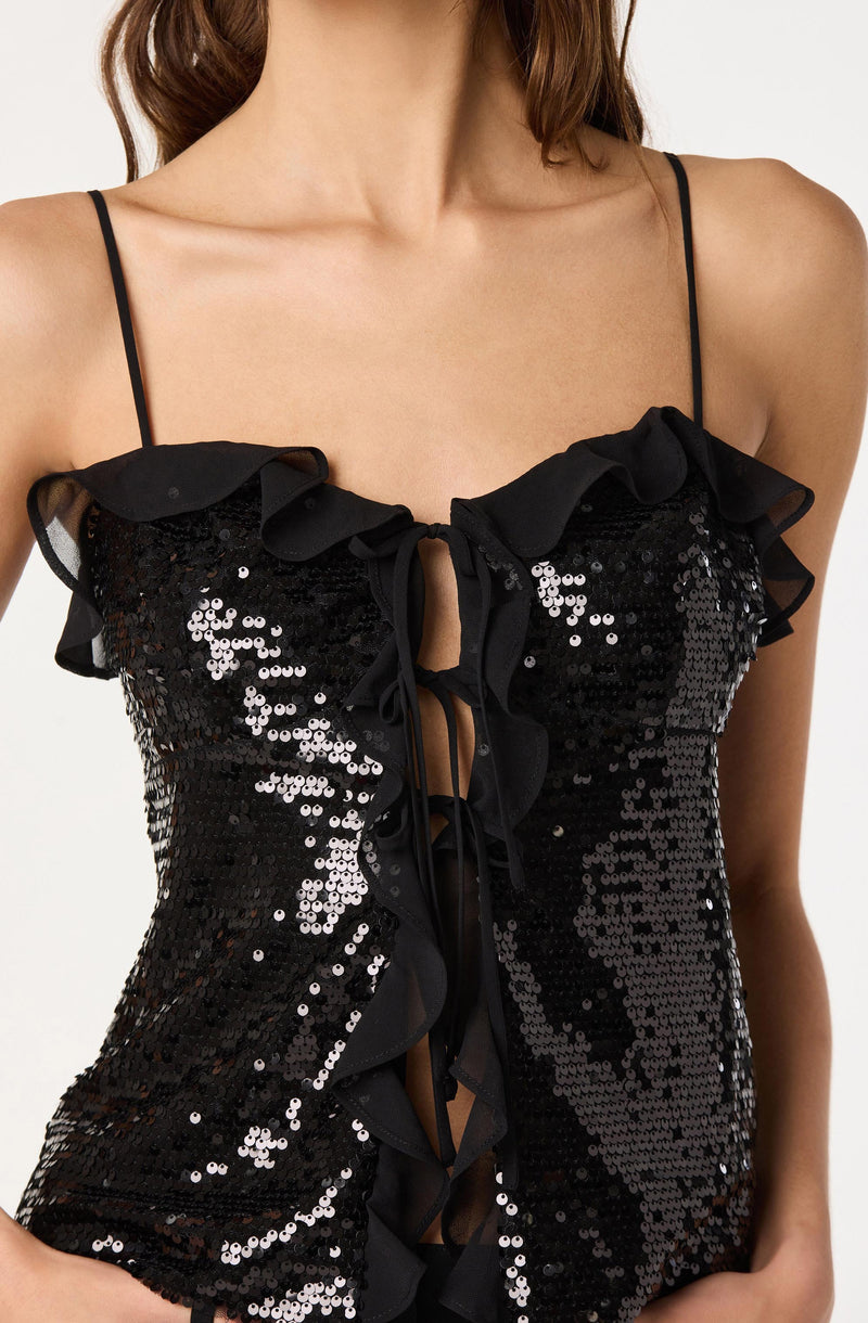 A woman is wearing a black sleeveless top with thin spaghetti straps. The top features a front detail with ruffles along the neckline and down the center, with a keyhole cutout at the top. The fabric is covered in shiny black sequins, giving it a sparkling appearance. The ruffles are made of a sheer, lightweight material that contrasts with the sequined body of the top.