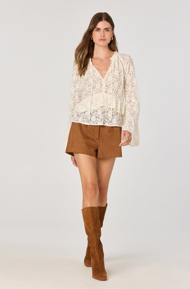 A woman is wearing a long-sleeved, cream-colored lace blouse with a V-neckline and a lace tie at the neck. She pairs it with high-waisted, brown shorts and knee-high brown suede boots.