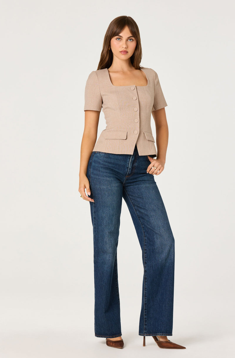 A woman is wearing a beige, short-sleeved blazer with a square neckline and a row of buttons down the front. The blazer features two small flap pockets at the waist. She pairs it with high-waisted, wide-leg blue jeans and brown pointed-toe heels.
