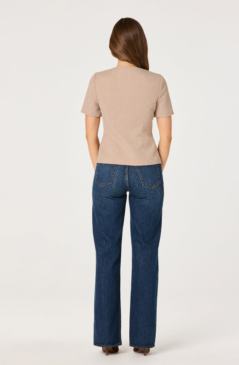 A woman is wearing a beige short-sleeve blazer with a fitted waist and structured shoulders, paired with high-waisted, straight-leg blue jeans.