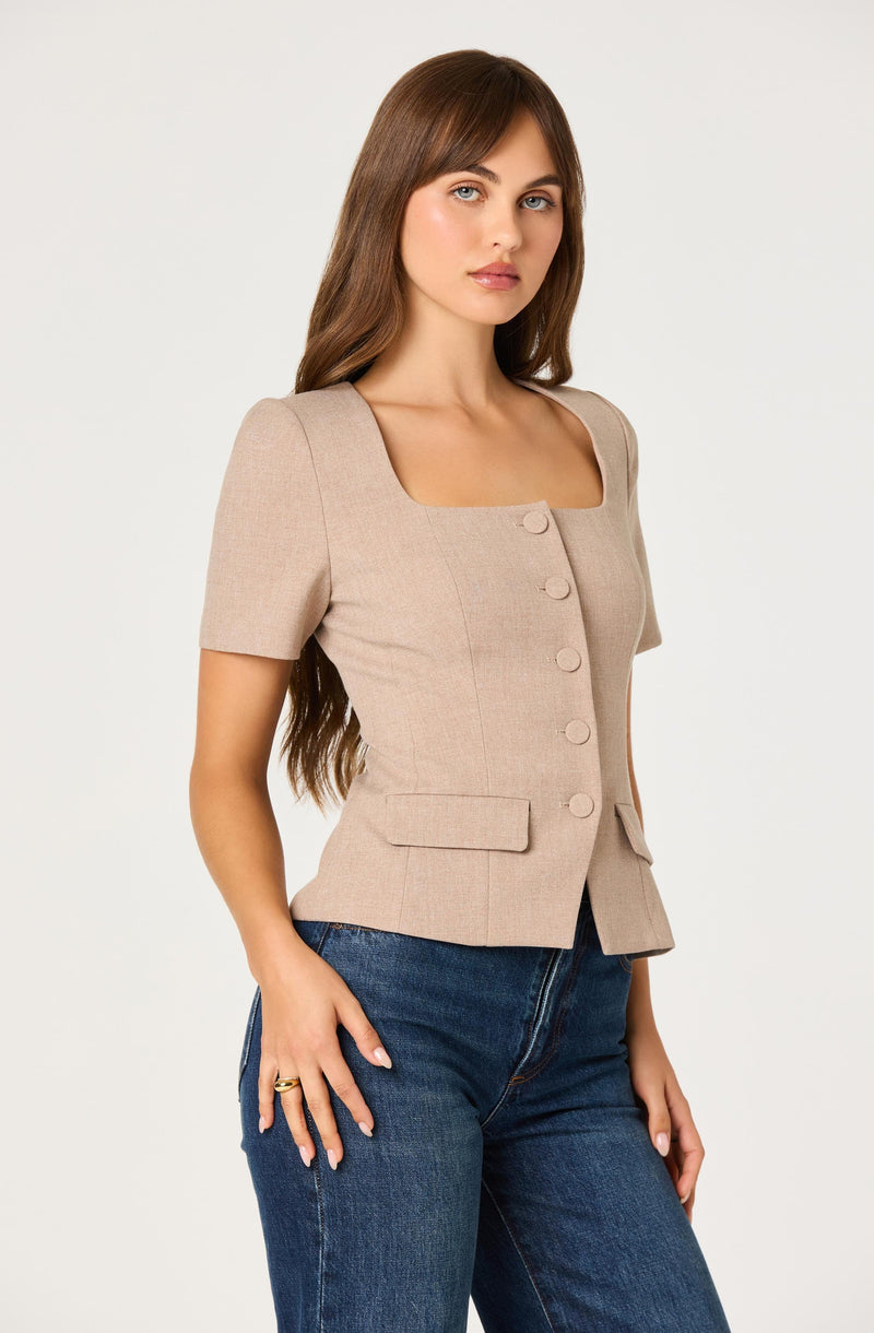 A woman is wearing a fitted, beige, short-sleeve blazer with a square neckline and large buttons down the front. The blazer features two flap pockets at the waist. She pairs it with dark blue jeans.