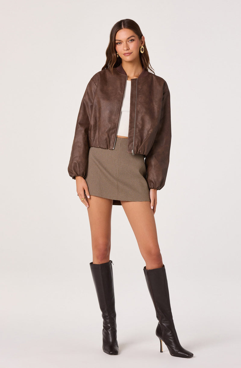 A woman is wearing a brown leather bomber jacket with a zipper front and puffed long sleeves, paired with a white ribbed crop top and a high-waisted, short, brown checkered skirt. She has accessorized with large circular earrings and is wearing tall black knee-high boots with high heels.