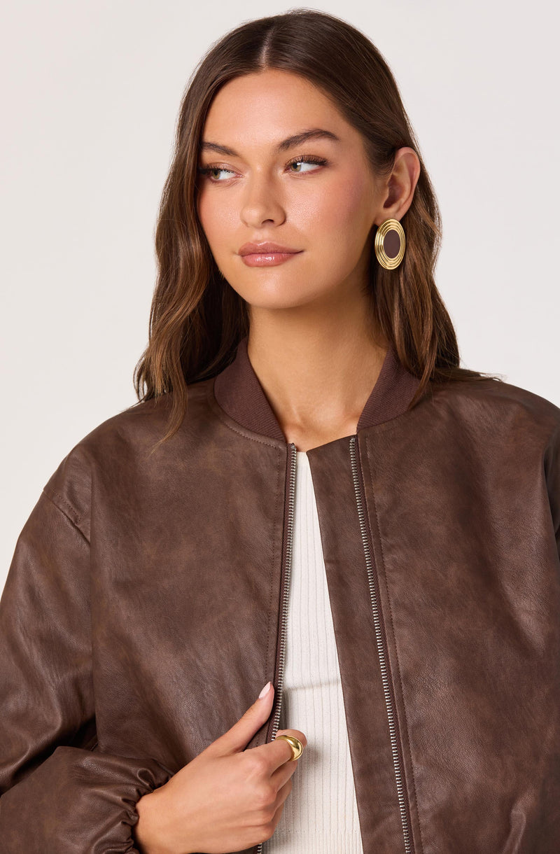 A woman is wearing a brown leather bomber jacket with a ribbed collar, paired with a white ribbed top underneath. She has large circular gold earrings and is also wearing a gold ring on her finger.
