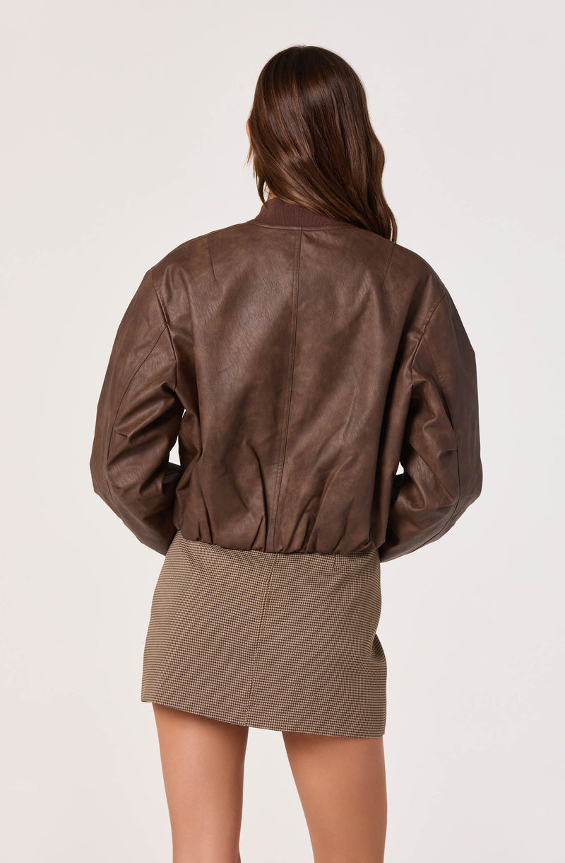 A woman is wearing a brown leather bomber jacket with a relaxed fit and ribbed collar, cuffs, and hem. She is also wearing a high-waisted, fitted mini skirt with a small checkered pattern in neutral tones.