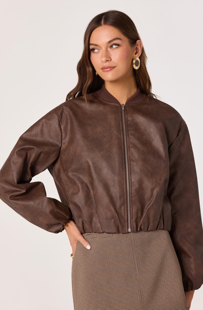 A woman is wearing a brown leather zip-up jacket with a stand-up collar and elasticized cuffs. She is also wearing gold hoop earrings and a high-waisted skirt with a small, checkered pattern.
