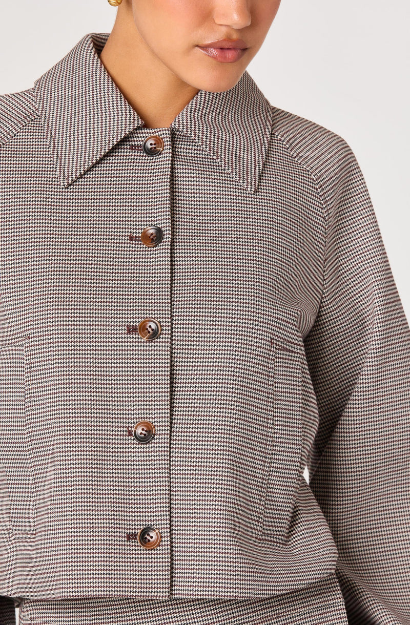 A woman wearing a long-sleeved button-up shirt with a small checkered pattern in shades of brown, black, and white. The shirt features a pointed collar and brown buttons down the front.