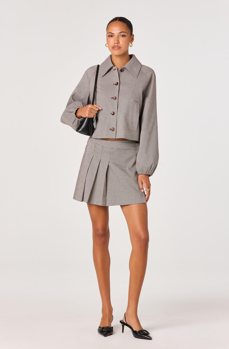 A woman is wearing a gray cropped jacket with a collar and large buttons, paired with a matching high-waisted pleated mini skirt. The outfit features a subtle checkered pattern. She accessorizes with gold hoop earrings, rings, and black pointed-toe heels with ankle straps. She carries a small black shoulder bag.