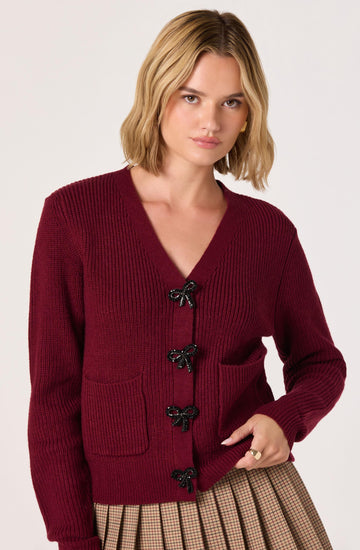 A woman is wearing a deep red, knitted cardigan with a V-neckline. The cardigan has black bow-shaped buttons down the front and a front pocket. She pairs it with a plaid skirt in shades of brown and beige.