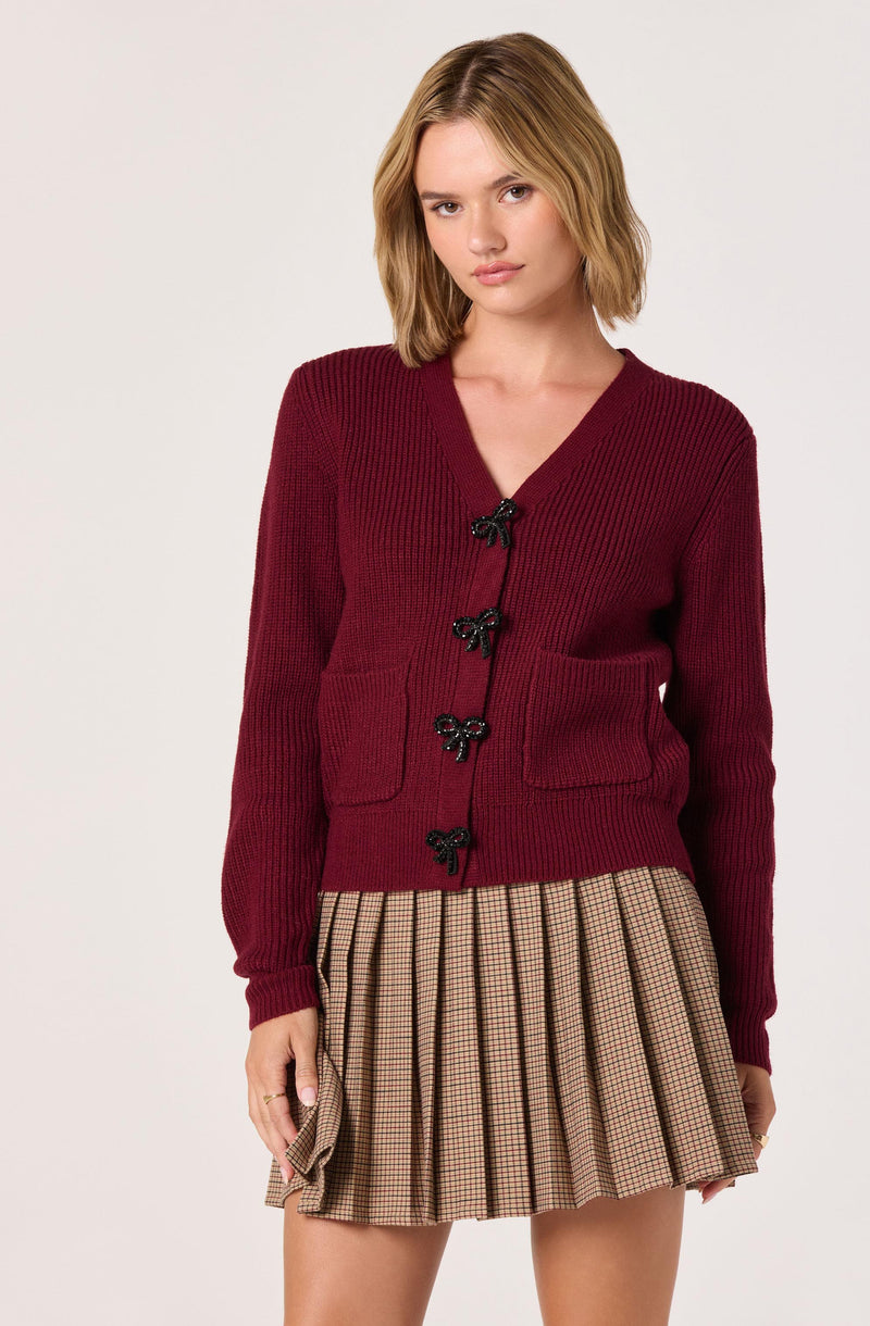 A young woman is wearing a burgundy knit cardigan with black bow-shaped buttons and two front pockets. She is also wearing a tan plaid pleated skirt with thin black and red lines.